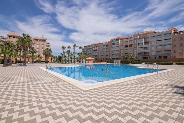A swimming pool with steps, surrounded by a patterned walkway, and adjacent to a multi-story building with balconies.