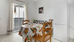 A dining area with a table covered in a fruit-patterned tablecloth, wooden chairs, a white refrigerator, a potted plant, and a window with curtains.