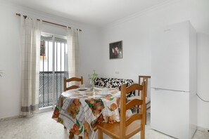Comedor con una mesa cubierta por un mantel estampado con motivos de frutas, sillas de madera, un refrigerador blanco, una planta en maceta y una ventana con cortinas.