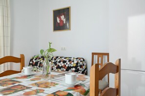 A dining table with a fruit-themed tablecloth, two white cups, a vase with a plant, and a polka-dotted cushion.