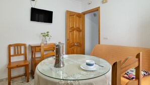 A room with a round glass table, a wooden chair, a sofa, a TV mounted on the wall, and a wooden door.