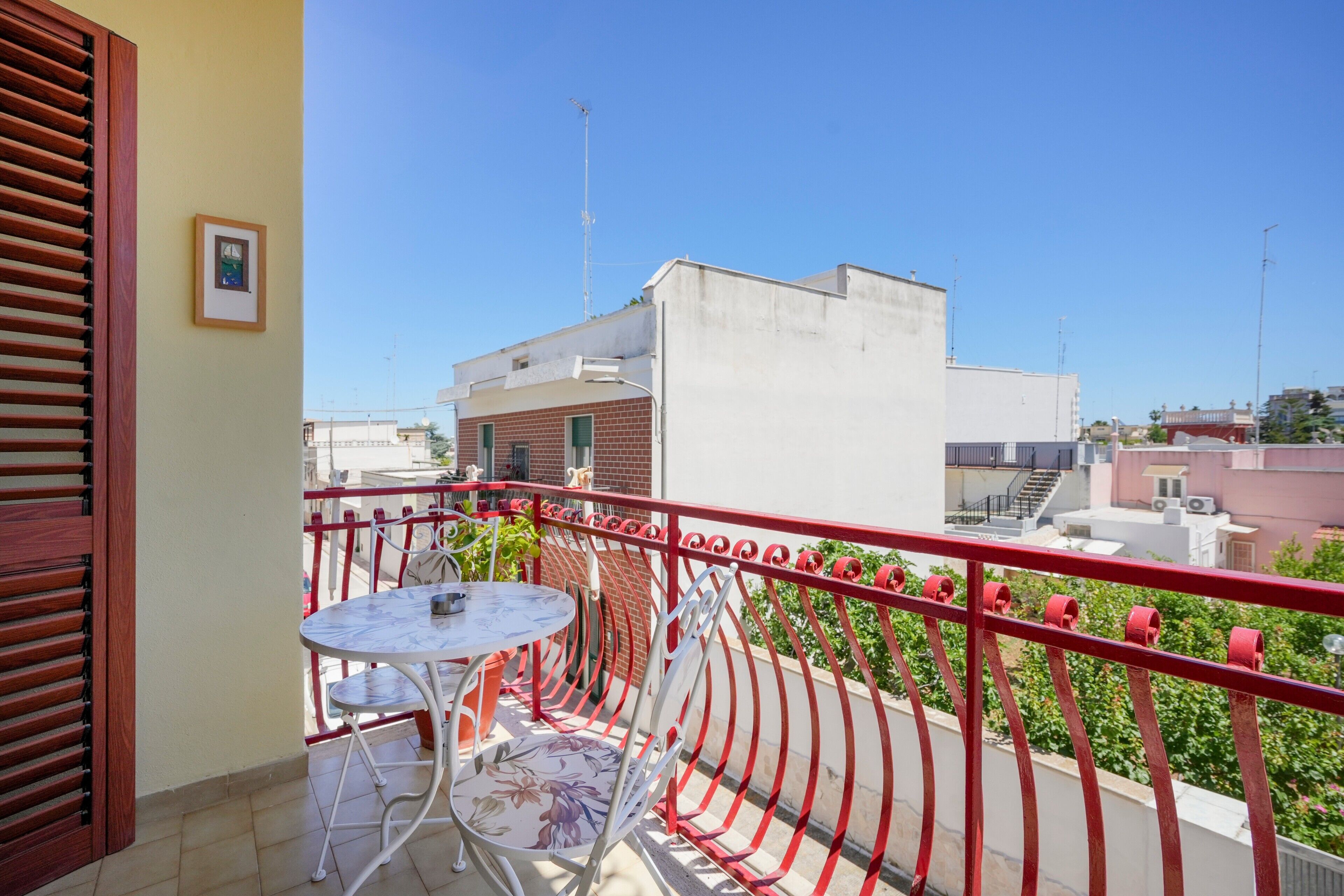 Un balcón con mesa y sillas, barandilla roja y vista a edificios.