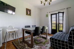 Un coin repas comprenant une table, des chaises, une chaise haute, un téléviseur, un lustre, une porte avec des rideaux et une vue à travers la porte.