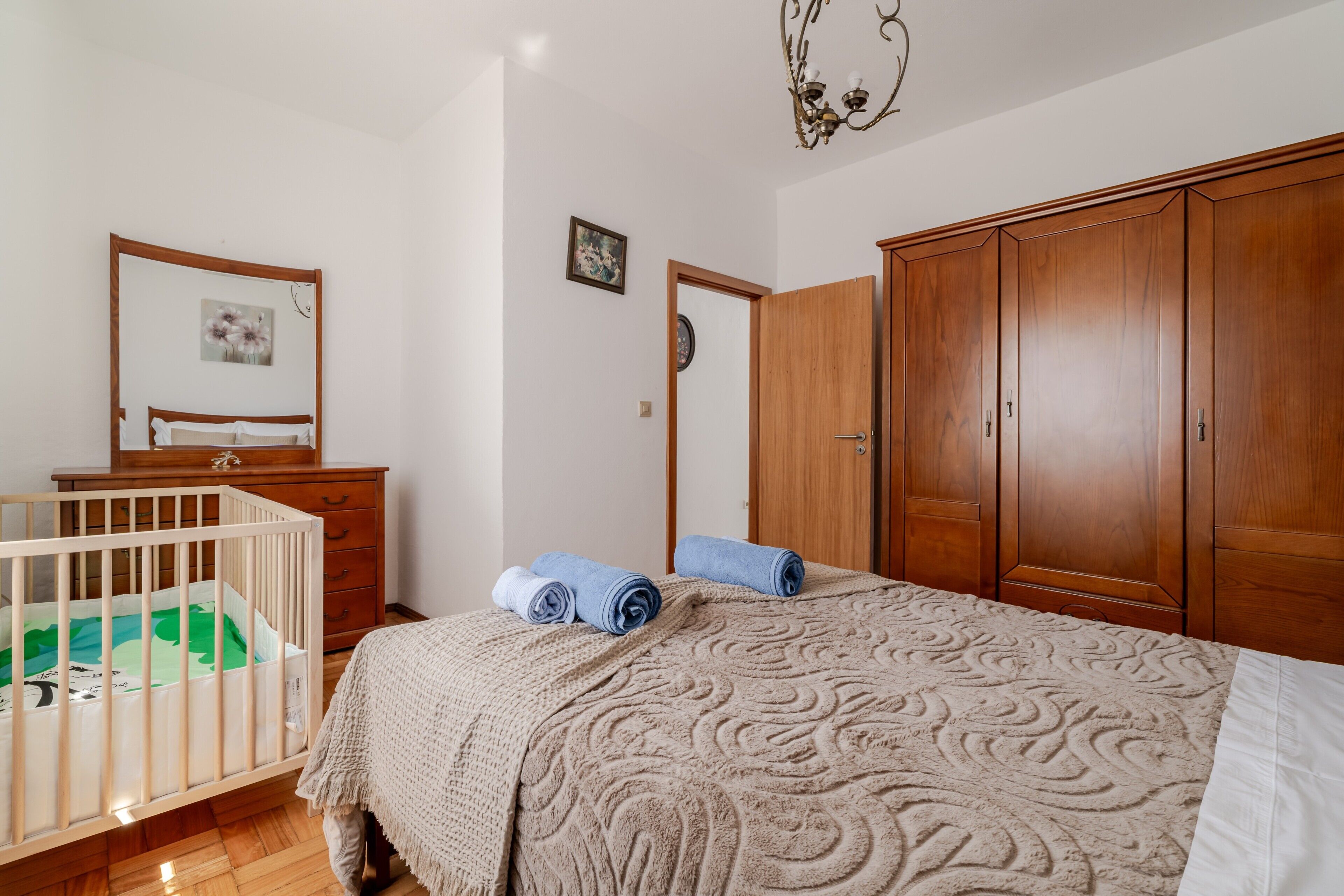 Ein Schlafzimmer mit einem Babybett, einem Holzschrank, einem Bett mit gemustertem Überwurf und einem Wandbild.
