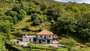 A two-story house with a tiled roof, surrounded by lush greenery and hills. - Superior Apartment with Mountain View, Shared Garden, and Wi-Fi Access (Ponte da Barca)