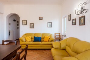 A living room with a yellow sofa, a wooden dining table, and chairs. There are framed pictures on the walls and a chandelier.