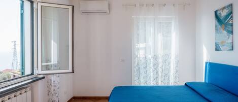 A bedroom with a large window, a radiator, a bed with blue bedding, and a curtain with a floral pattern.