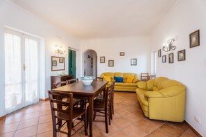 A living room with a dining table, yellow sofa, and a kitchen area visible through an archway.