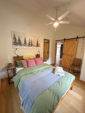 Une chambre à coucher avec un lit, un tableau représentant des arbres, un ventilateur de plafond et une porte en bois.