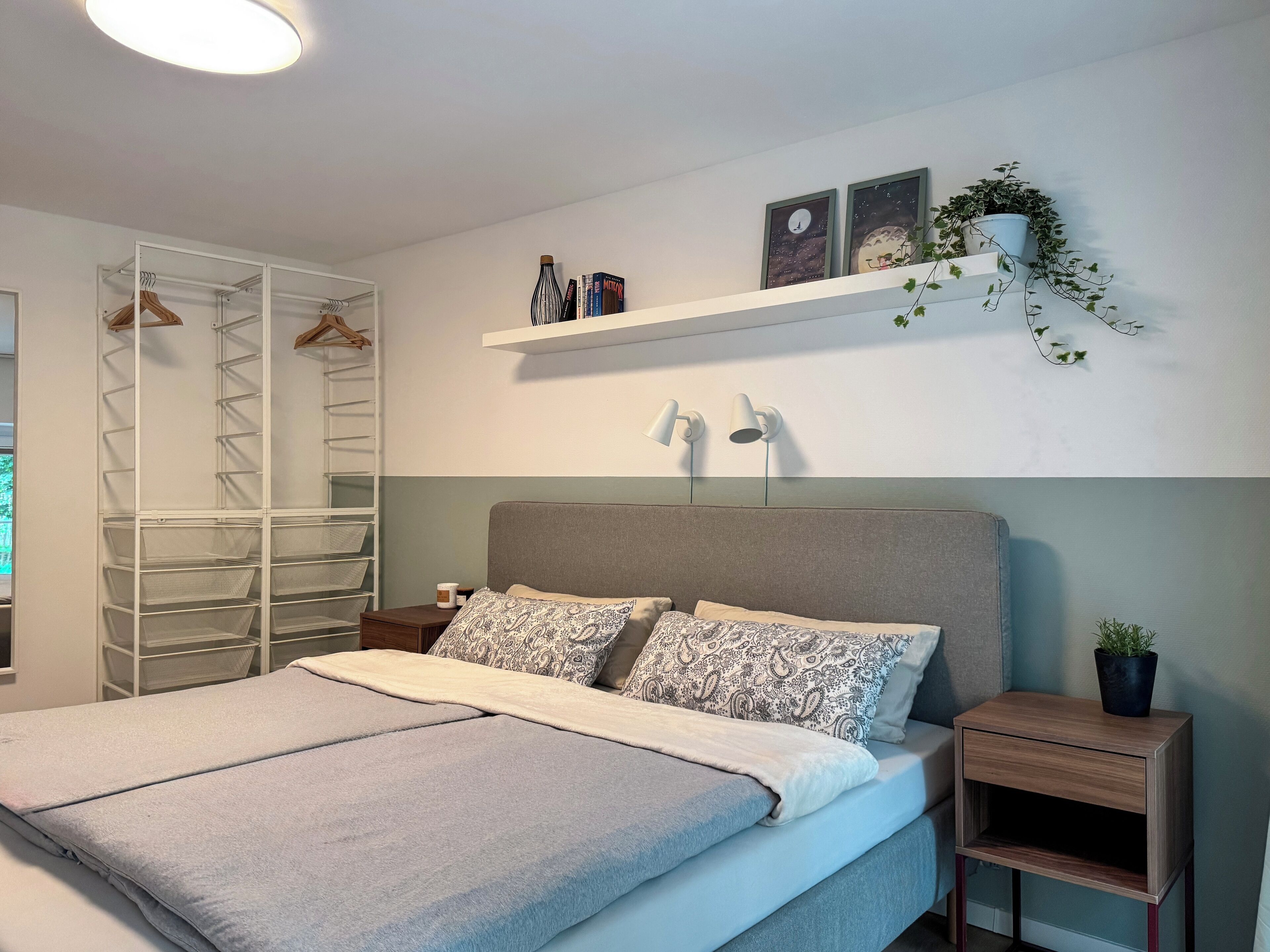 Une chambre bien rangée, avec un lit, des tables de chevet, une armoire et une étagère où sont disposés des objets décoratifs et des plantes.