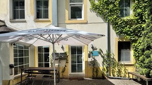 An outdoor patio area with a table and chairs under a large umbrella, a bench, and a building with multiple windows and ivy.