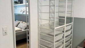 A room with a white metal shelving unit with multiple wire baskets, a mirror, and a bed with patterned pillows.