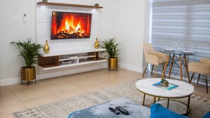 A modern living room with a flat-screen TV mounted on a white wall, a glass coffee table, and two beige armchairs.