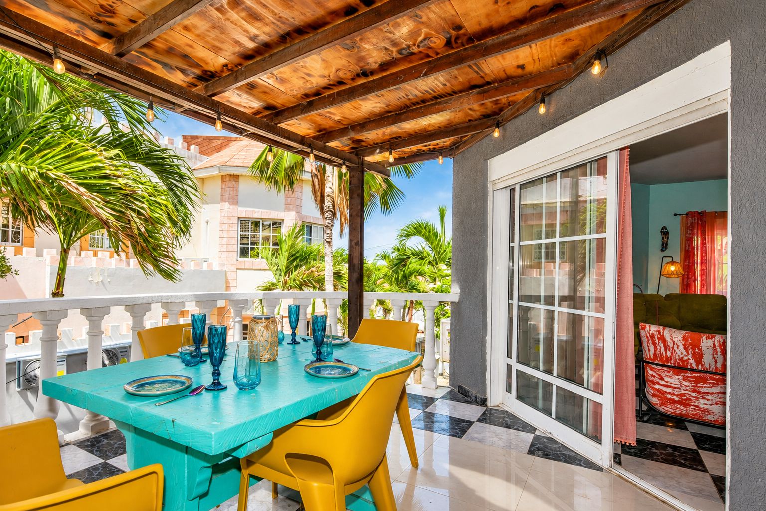 An outdoor patio with a wooden ceiling, a table set with blue glassware, yellow chairs, and a view of a building with a balcony.