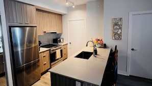 A modern kitchen with a stainless steel refrigerator, wooden cabinets, a built-in oven, and a large island with a sink. - Luxury 1BR Condo | Sophisticated & Stylish Stay by Square One (Mississauga)