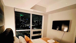 A hotel room with a bed, pillows, a TV mounted on the wall, and a window with a city view. - Luxury 1BR Condo | Sophisticated & Stylish Stay by Square One (Mississauga)