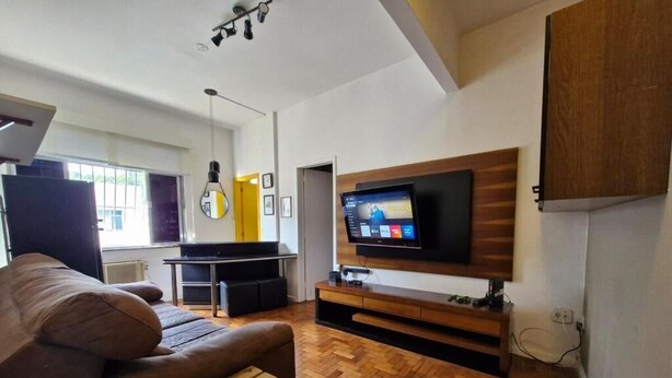A modern living room with a wooden herringbone floor, a brown sofa, a wooden bench, a flat-screen TV mounted on a wooden wall, and a kitchen area in the background. - SUH Copacabana 89 Beach View (Rio de Janeiro)