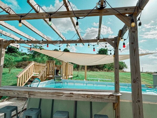 Una terraza de madera con piscina, una escalera de madera y una pérgola con luces colgantes.
