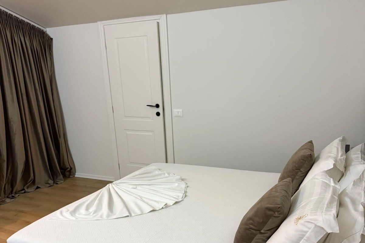 Een enkel bed met een witte matras en een houten hoofdeinde. Er is een deur met een handgreep en een gordijn dat gedeeltelijk is opengetrokken.