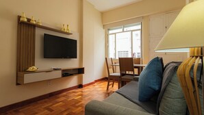 A living room with a flat-screen TV mounted on a wooden shelf, a sofa with cushions, a dining table with chairs, and a large window with a view of buildings. - SUH Copacabana 76  (Rio de Janeiro)