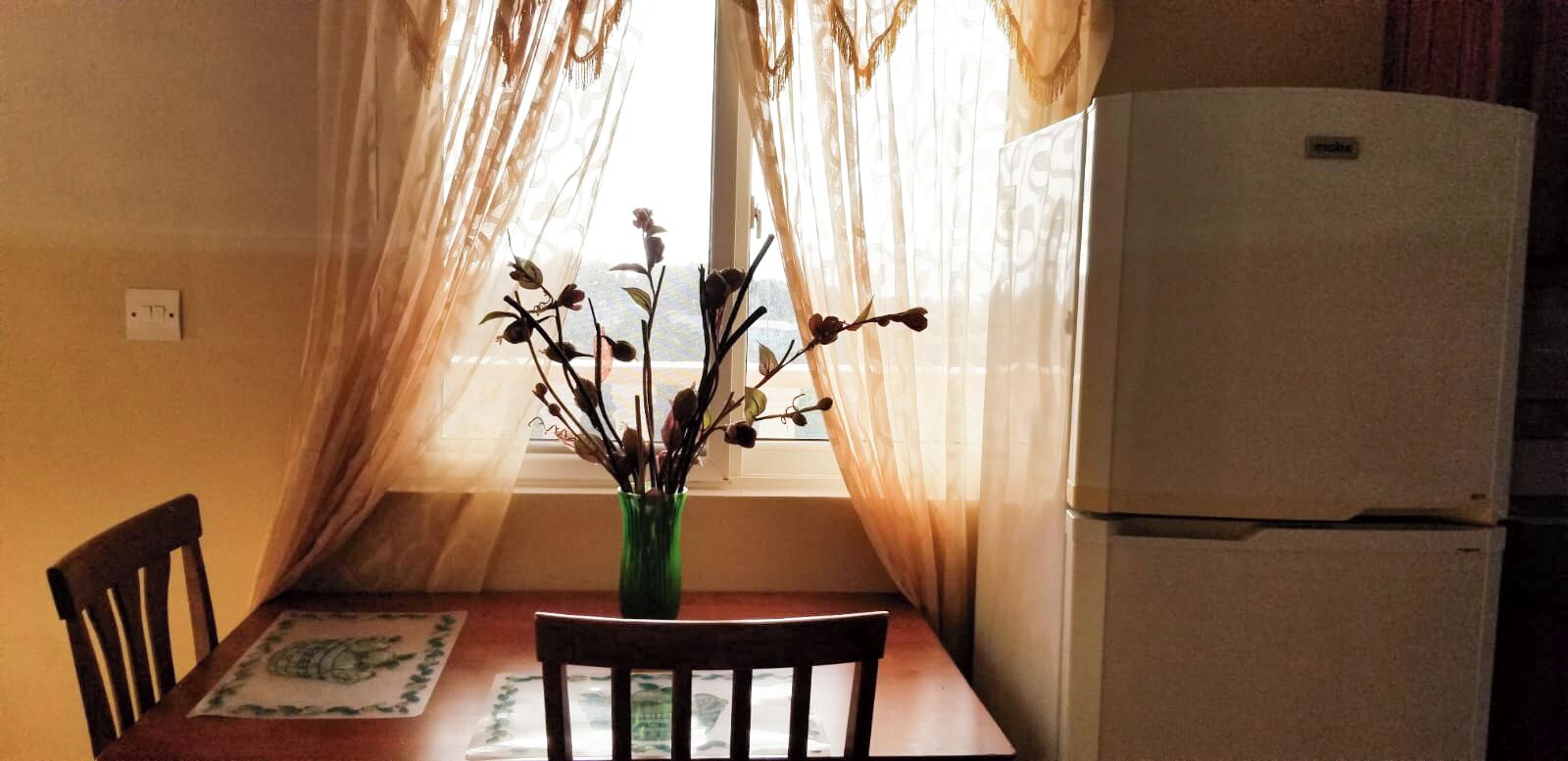 A kitchen with a table, chairs, a refrigerator, and a window with sheer curtains.