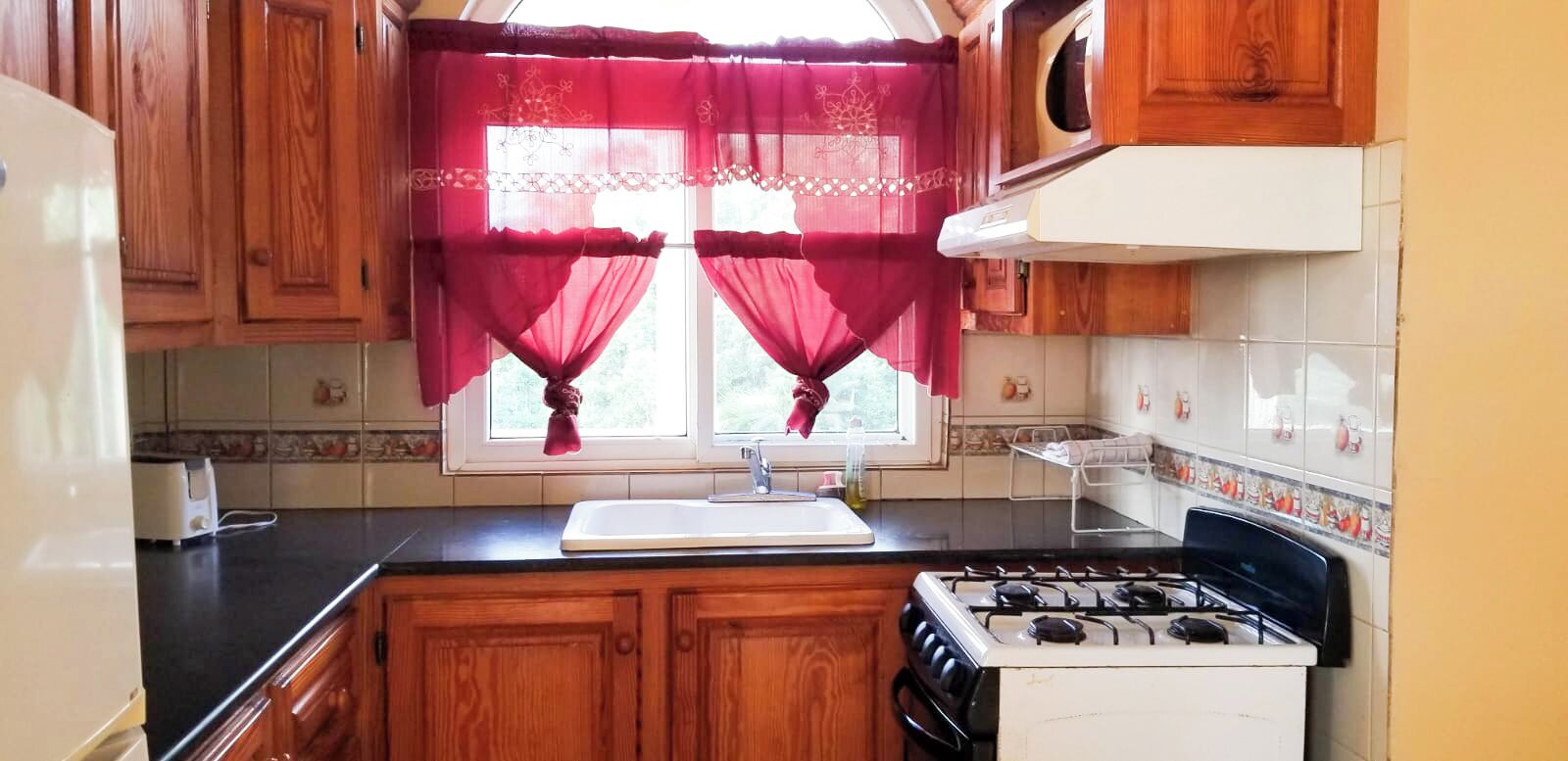 Cocina compacta con gabinetes de madera, estufa a gas, lavamanos y una ventana con cortinas rojas.