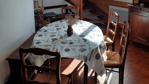 A dining area with a round table covered by a tablecloth with a leaf pattern, surrounded by wooden chairs. A wooden staircase with a railing is visible in the background, leading to an upper level.