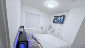 Une chambre à coucher avec un lit, un bureau avec une lampe, un téléviseur fixé au mur et une fenêtre.