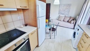 A compact kitchen with a stove, oven, and washing machine, a dining area with a glass table and chairs, and a sofa in the background. - Kainon Asti (Lefkada)