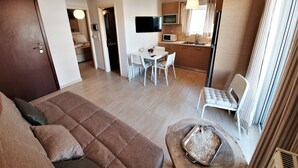A modern living room with a sofa, a dining area with a table and chairs, a kitchen with a TV, and a large window with curtains. - Kainon Asti (Lefkada)