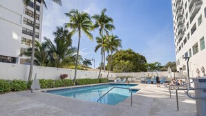 Piscina con palmeras, sillas de descanso y un edificio blanco.