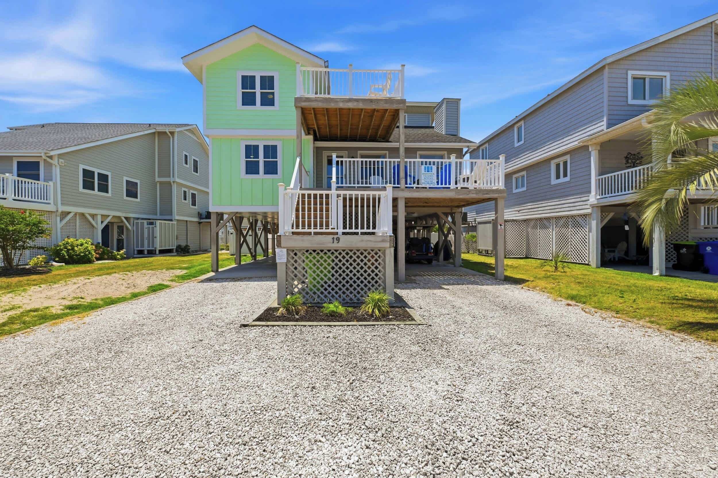 A multi-story residential building with a covered front porch, a small garden area, and a gravel driveway.