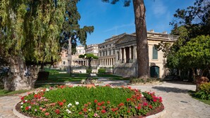A courtyard with a central flower bed, a large tree, and a classical building with columns. - Anthia: Balcony and BBQ, 3 min to Airport, WiFi, Aircon (Kanoni)
