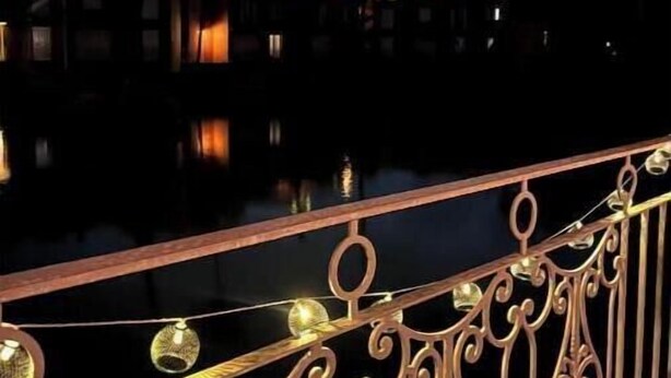 Vista nocturna de un paisaje urbano con edificios y palmeras, superficie acuática reflectante y barandales de metal ornamentados con luces.