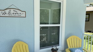 Un porche con dos sillas Adirondack amarillas, una mesita y una ventana con persianas. Un cartel de bienvenida está colgado en la pared.