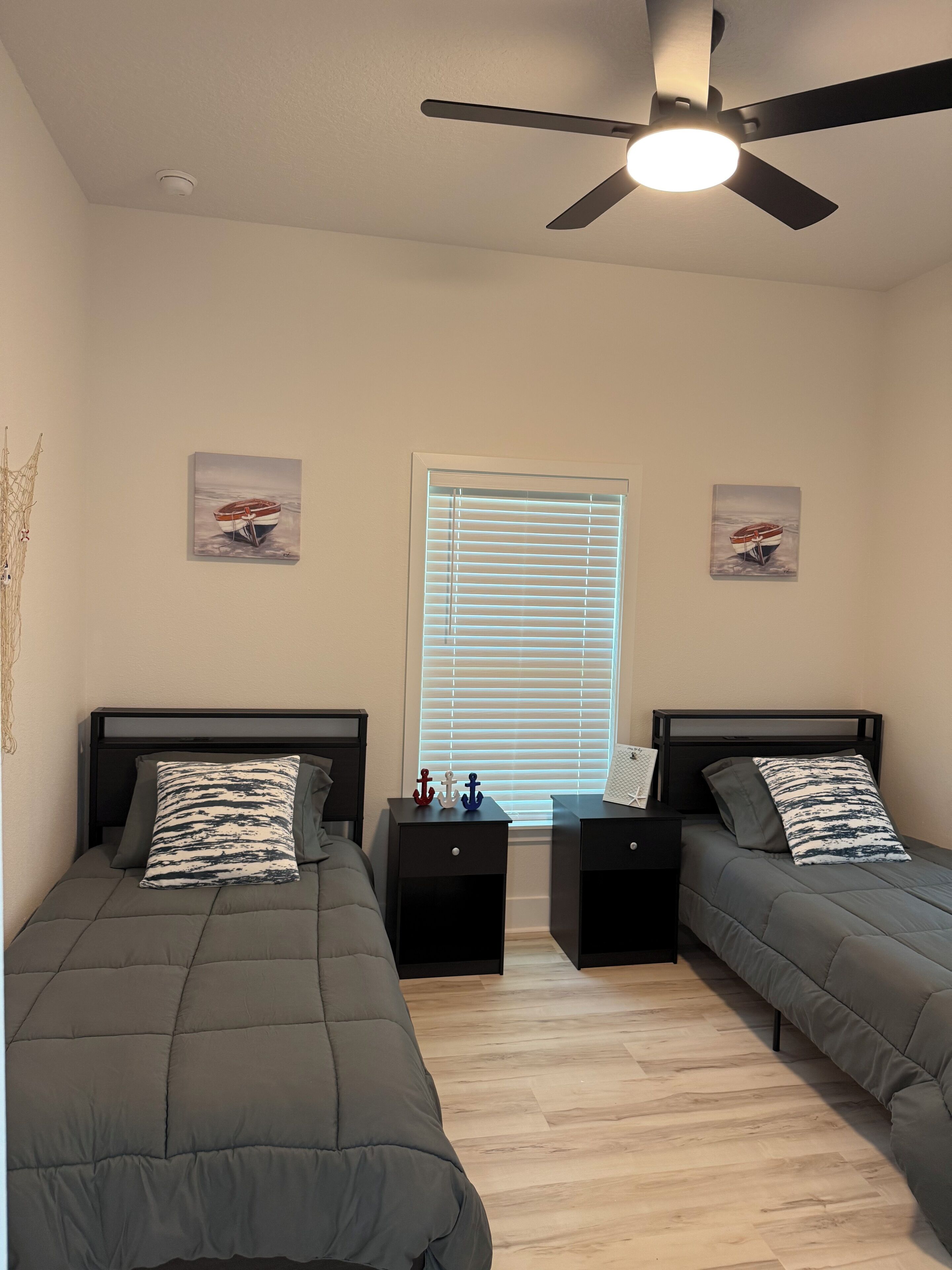 A bedroom with two beds, a ceiling fan, a window with blinds, and two nightstands with decorative items.