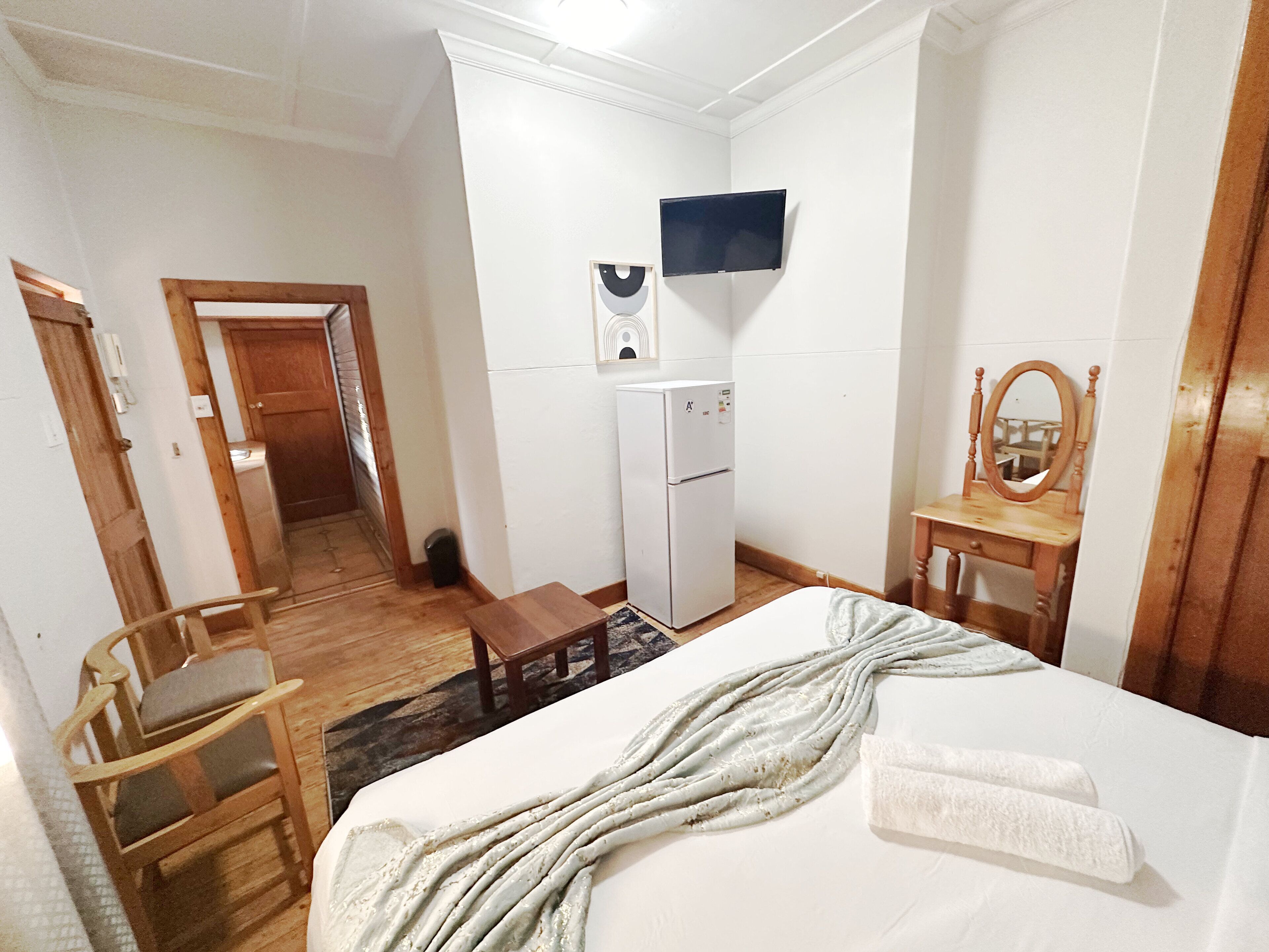 Un dormitorio con una cama, una silla de madera, una mesita, un espejo, un televisor y un refrigerador.