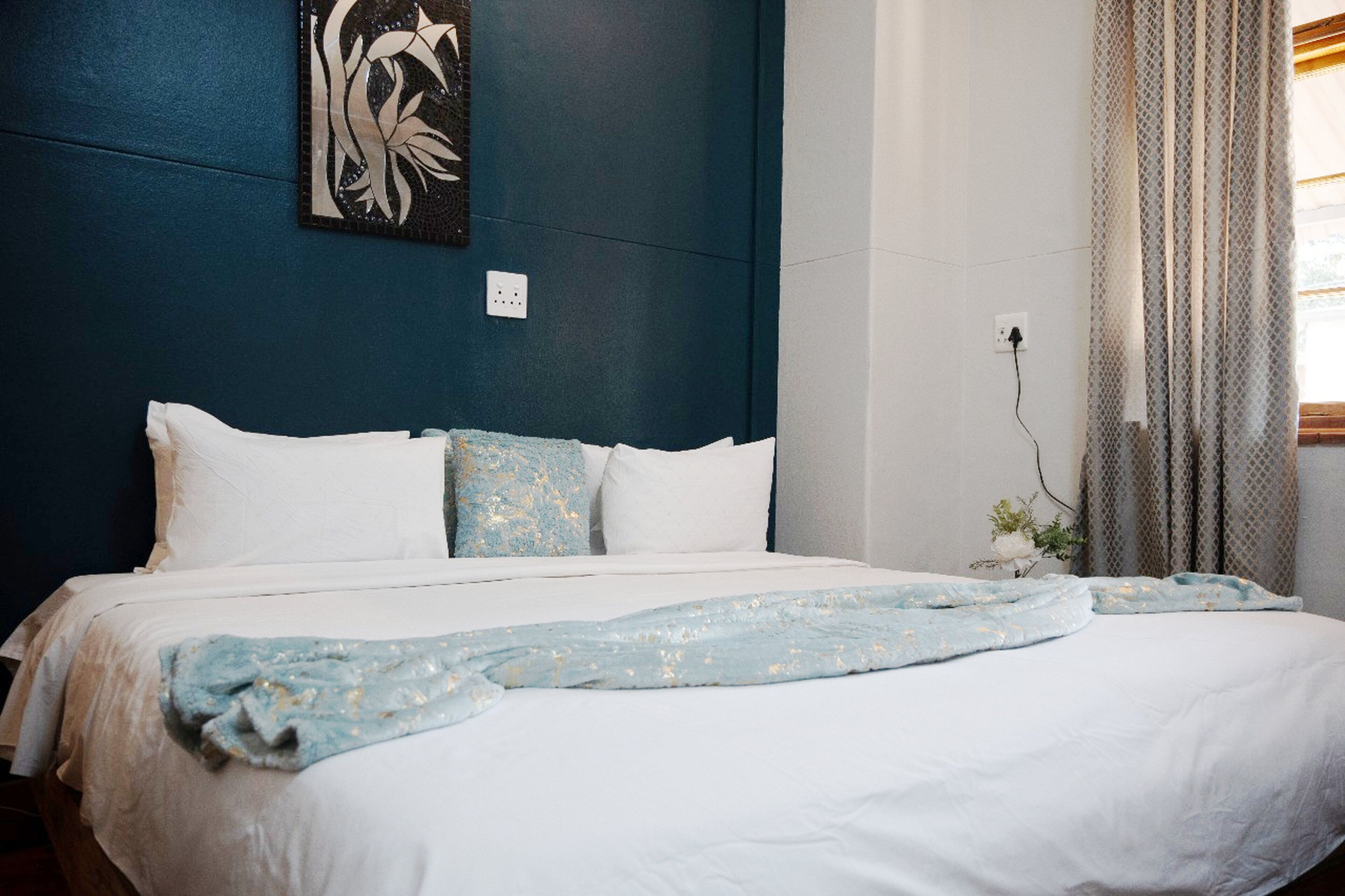 A neatly made bed with white and light blue bedding, a wall-mounted power outlet, and a window with patterned curtains.