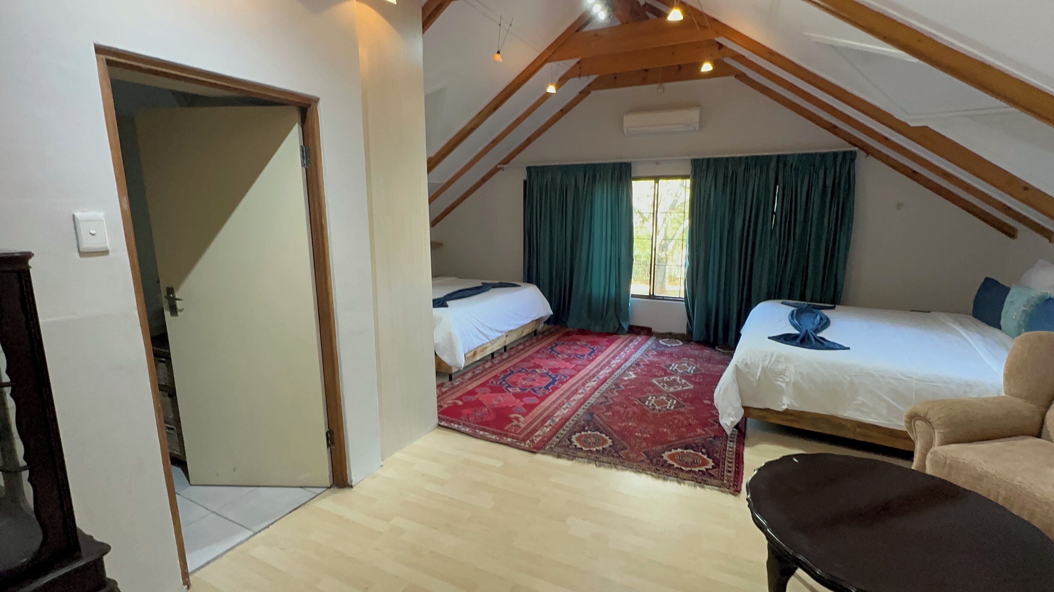 A room with two beds, a red patterned rug, wooden beams, and a large window with curtains.