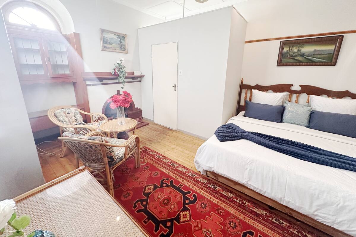 Une chambre à coucher avec un lit, une chaise, une petite table sur laquelle se trouve un vase, un tapis et un tableau accroché au mur.