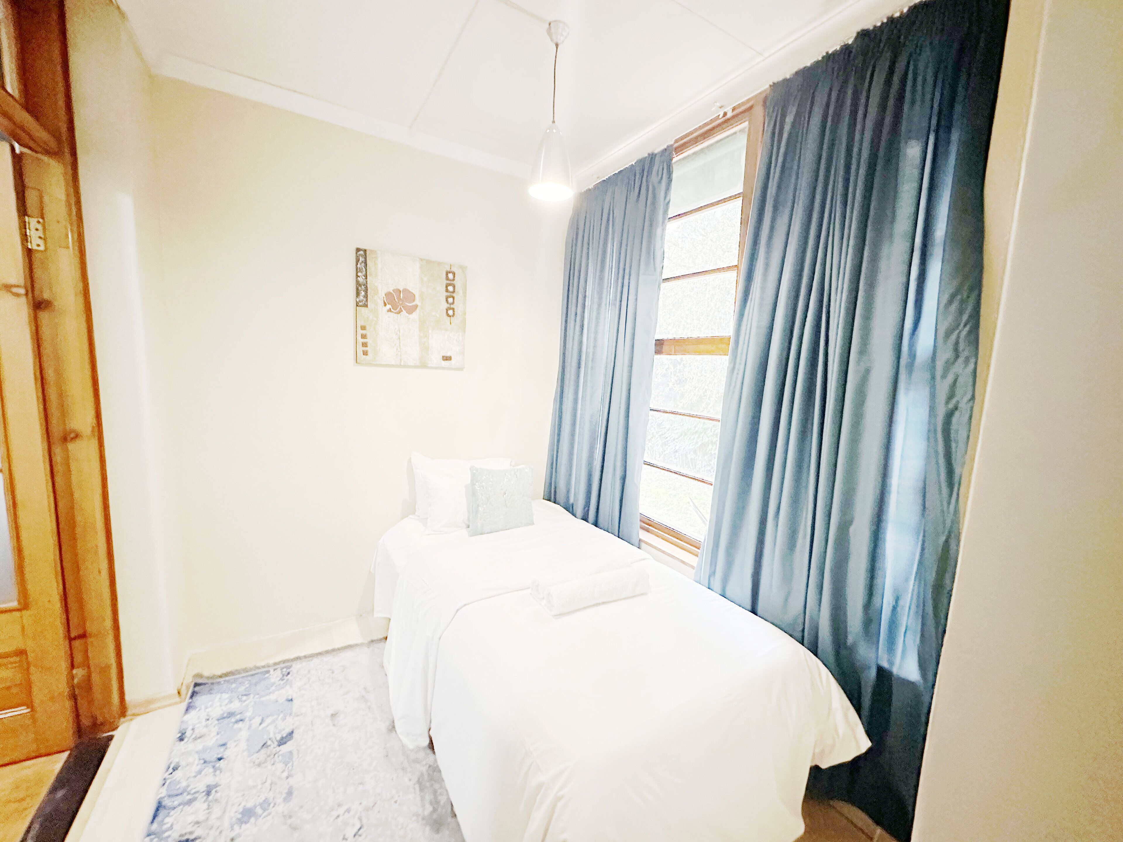A bedroom with a bed, a nightstand, a window with curtains, and a wall-mounted artwork.