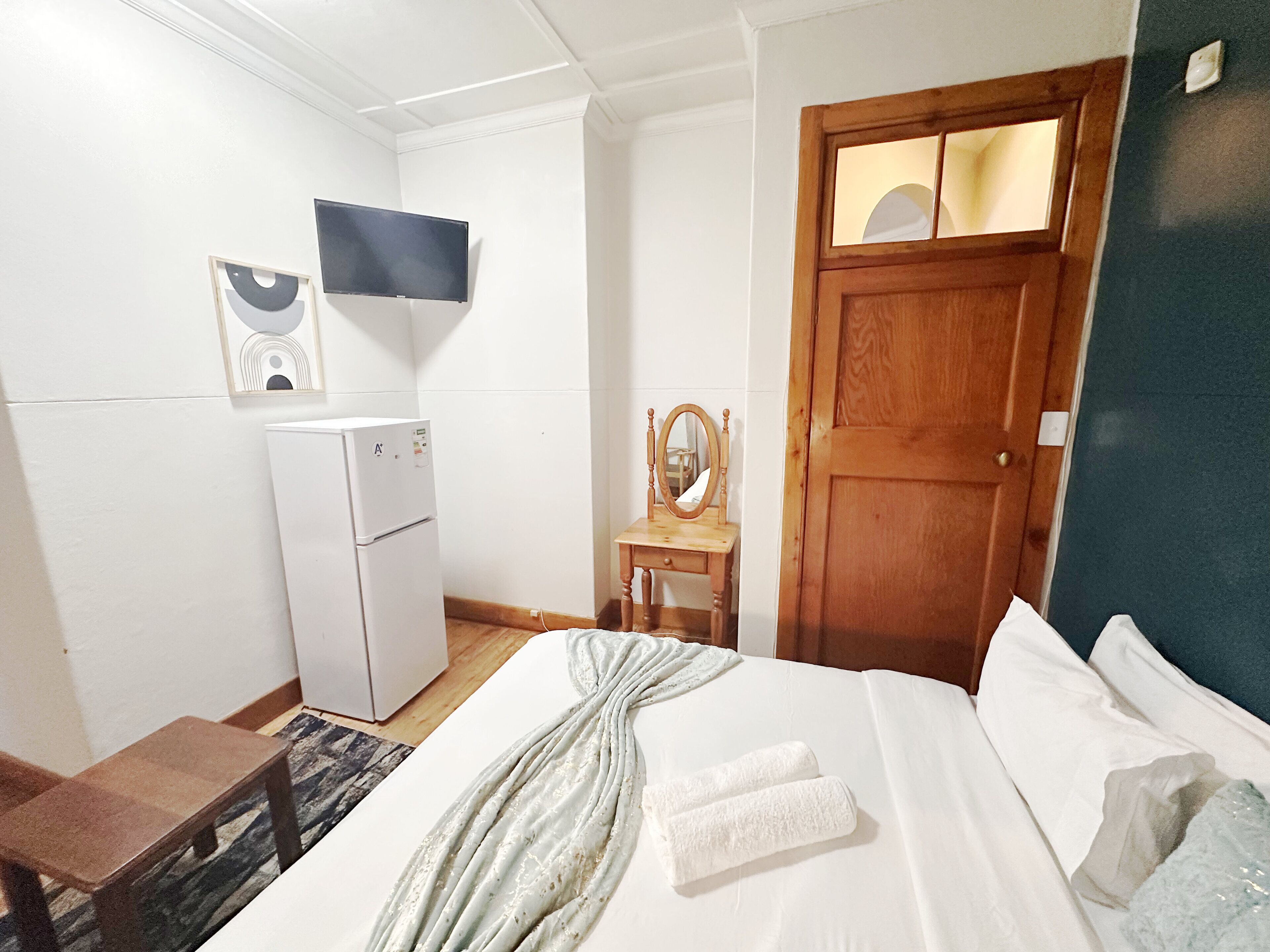 A hotel room with a bed, a wooden door, a small table with a mirror, a white refrigerator, and a wall-mounted TV.