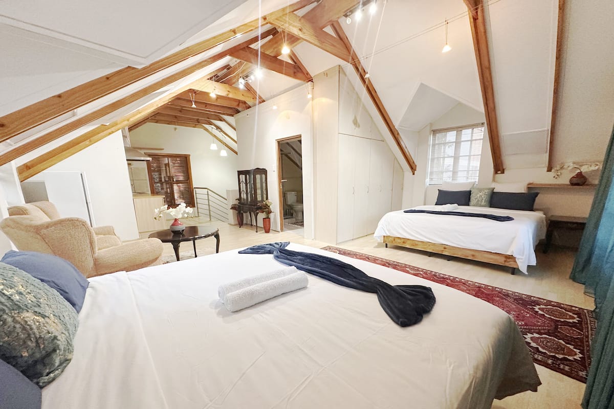 Une chambre avec un grand lit, un lit plus petit et un canapé. Le plafond est en bois et un escalier mène à une autre pièce.