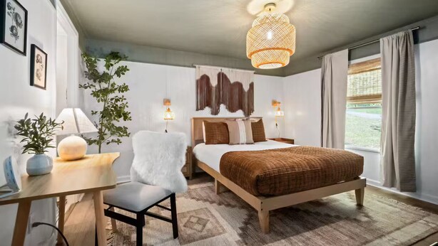 A bedroom with a bed, bedside tables, a desk, a chair, a hanging light fixture, and a window with curtains.