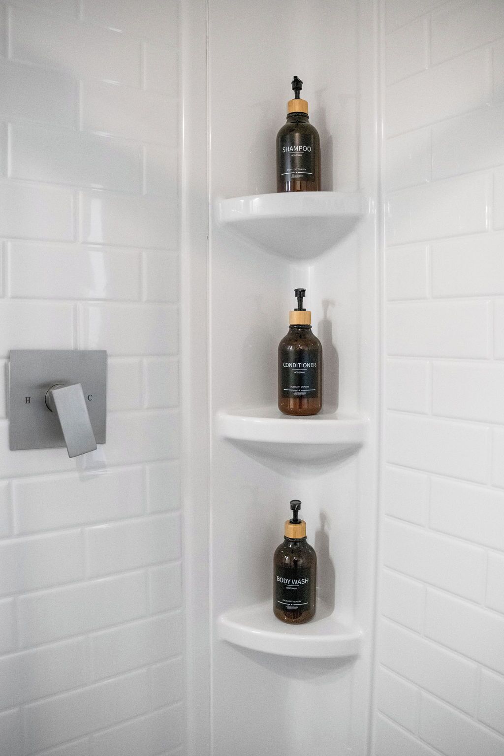 A bathroom with white tiled walls, a white sink, and three shelves holding bottles of shampoo, conditioner, and body wash.