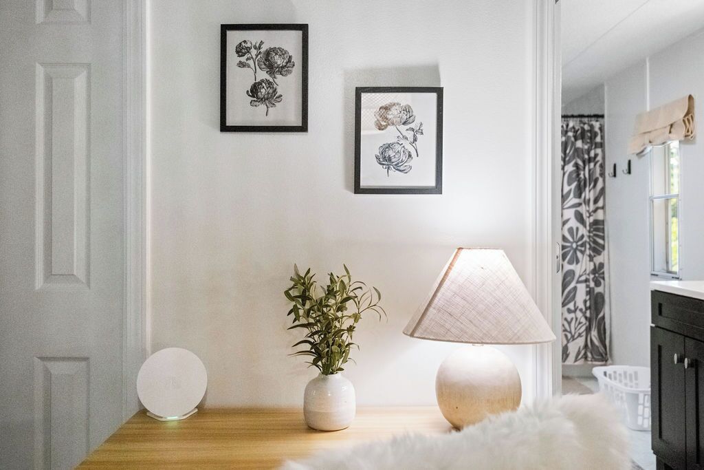 Une pièce avec une porte blanche, une table ronde blanche, un vase blanc contenant des plantes vertes, une lampe blanche et deux reproductions encadrées en noir et blanc représentant des motifs floraux, accrochées au mur.