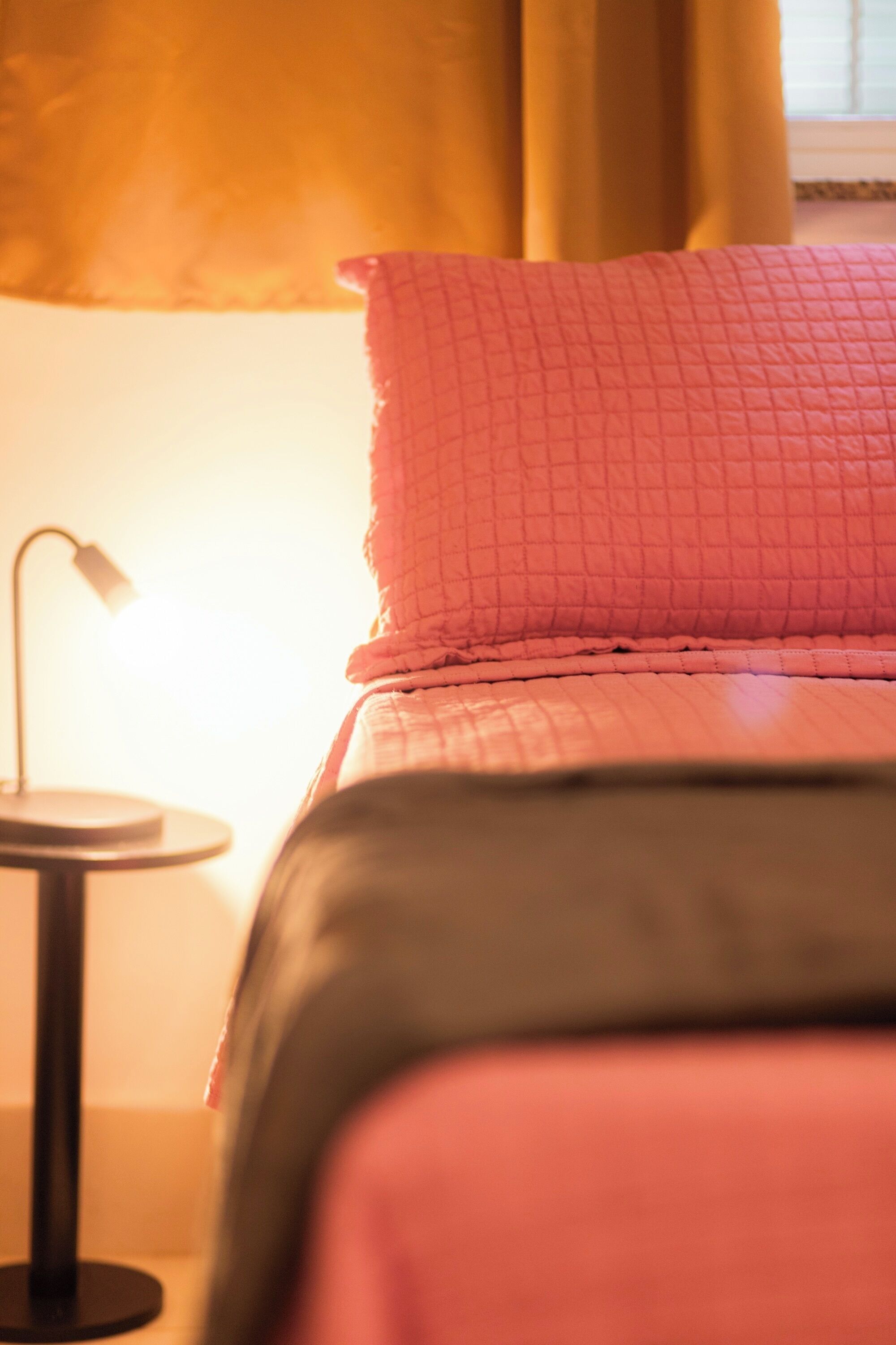 A bed with a quilted pink and black cover, a bedside lamp, and a window with curtains.