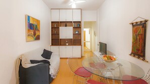 Un espacio habitable compacto con un sofá, una mesa de comedor de vidrio con sillas, un ventilador de techo y una estantería.
