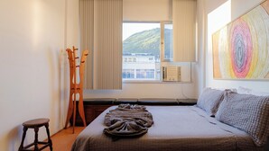 Un dormitorio con cama, una silla de madera, un taburete, una ventana con cortinas y un cuadro abstracto y colorido.