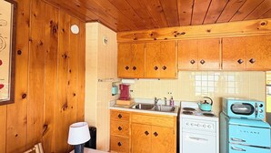 Un intérieur de chalet chaleureux avec des murs et un plafond en bois, un coin repas avec une table et des chaises, et une petite cuisine équipée d’appareils électroménagers d’époque.